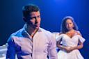 A man in a white dress shirt looks serious under blue stage lighting, while a woman in an off-shoulder white gown stands blurred in the background, suggesting a dramatic or emotional scene.