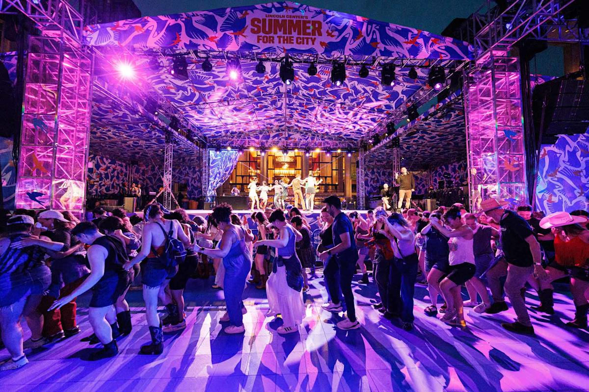 A vibrant outdoor stage event at night, with colorful lights and a “Summer for the City” banner, shows people dancing energetically in front of the stage under a decorative canopy.