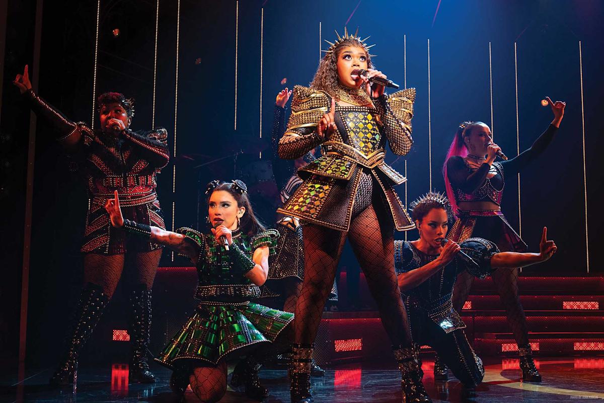 Five performers in ornate, studded costumes sing and strike dynamic poses on a colorful stage, with dramatic lighting and vertical light bars in the background.