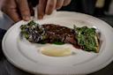 A chef’s hands plate grilled leafy greens and a glazed steak beside a swirl of creamy mashed potatoes on a white dish.