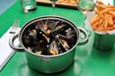 A pot of steamed mussels with herbs sits on a green table next to a cup of golden French fries, a glass of white wine, and a fork and napkin.