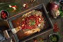 A whole cooked fish in a rectangular pan, topped with chopped red chilies, garlic, and green peppercorns, surrounded by fresh ingredients like chili peppers, red onion, and herbs on a dark wooden table.
