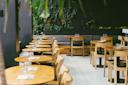 A modern, empty restaurant with wooden tables and chairs, black cushioned benches, glasses and napkins set on each table, and green plants decorating the dark walls.