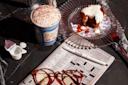 A crossword puzzle with a pen, glasses, and a Greek-style coffee cup filled with a foamy drink, sprinkled with cinnamon. A plastic creamer cup is open nearby. A slice of cake with icing rests on a glass plate.