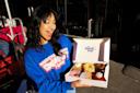 A smiling woman in a blue "Underground Donut Tour" sweatshirt holds an open box of assorted donuts from The Donut Pub while standing outside in sunlight.