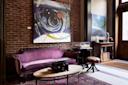 A cozy room with exposed brick walls, a purple vintage sofa, a wooden coffee table with a typewriter, abstract artwork on the wall, and eclectic decor including scientific objects and glass domes.