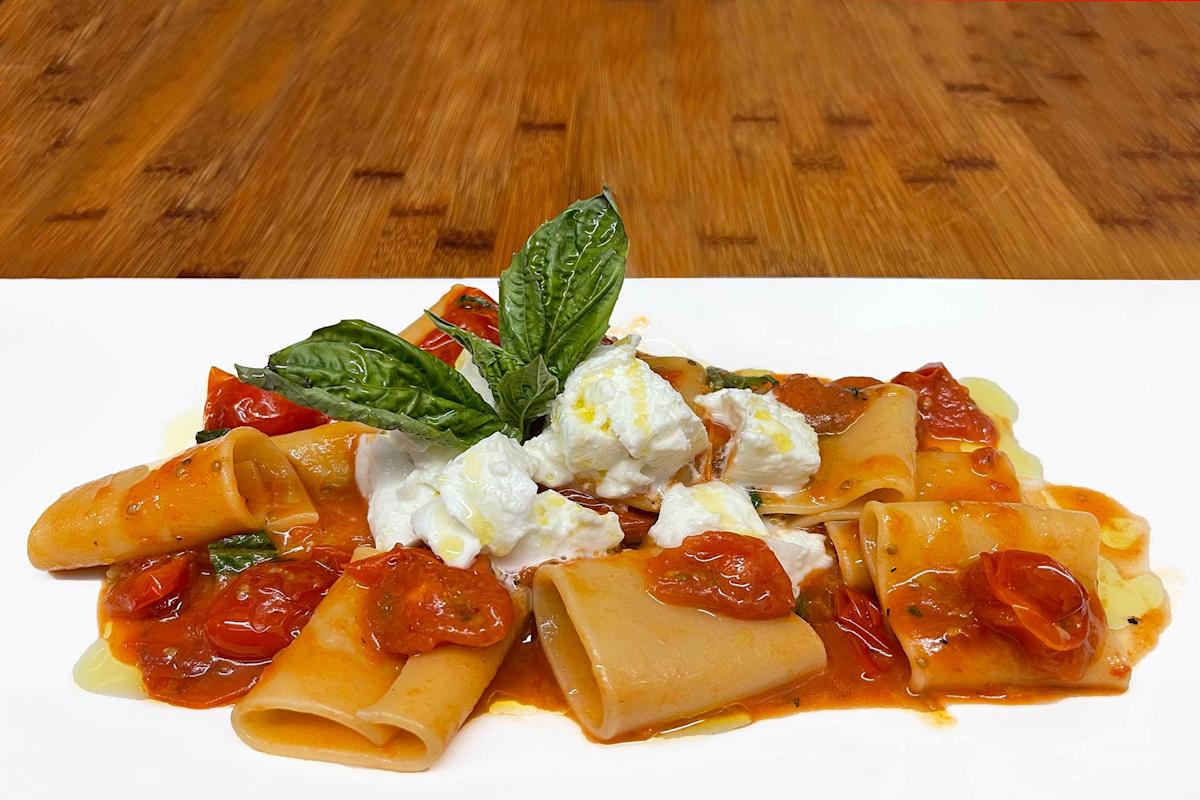 A plate of pasta with tube-shaped noodles, cherry tomato sauce, fresh basil leaves, and dollops of creamy cheese, served on a white plate with a wooden surface in the background.