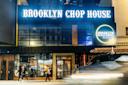 Street view of Brooklyn Chop House at night. The facade features large illuminated signage. Pedestrians walk on the sidewalk, and a car passes by in the foreground. A round sign advertises the rooftop bar.