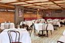 A cozy restaurant interior with brick walls, tiled floors, and wooden beam ceilings. Tables are covered with white cloths and surrounded by dark wooden chairs. Walls are adorned with black and white photographs and shelves of wine bottles.