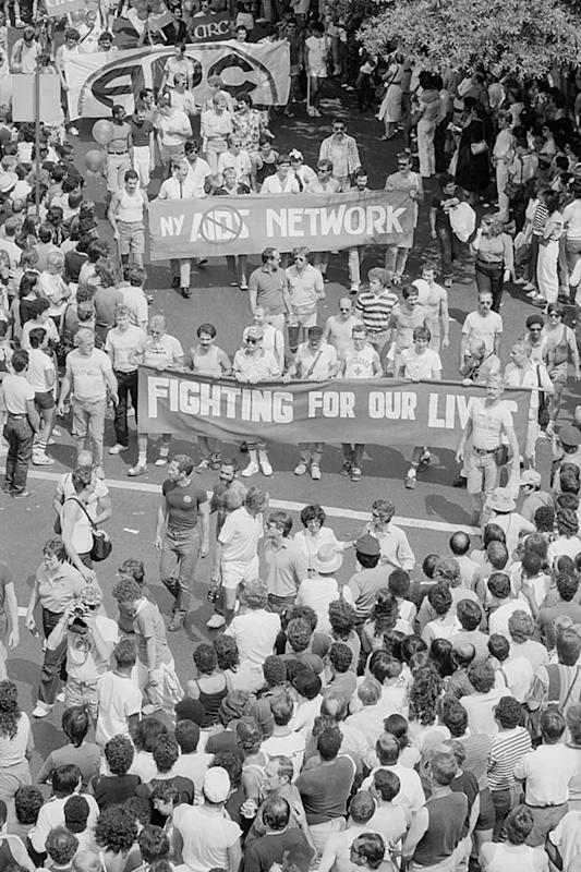 Black-and-white photo of a large crowd at a street protest. People hold banners reading “NY NETWORK” and “Fighting For Our Lives,” surrounded by onlookers, some of whom are clapping and wearing t-shirts.