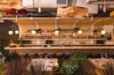 A stylish bar with hanging fringe lamps, round pendant lights, a long wooden counter with blue stools, and shelves stocked with bottles. Green plants decorate the space, creating a warm, inviting atmosphere.