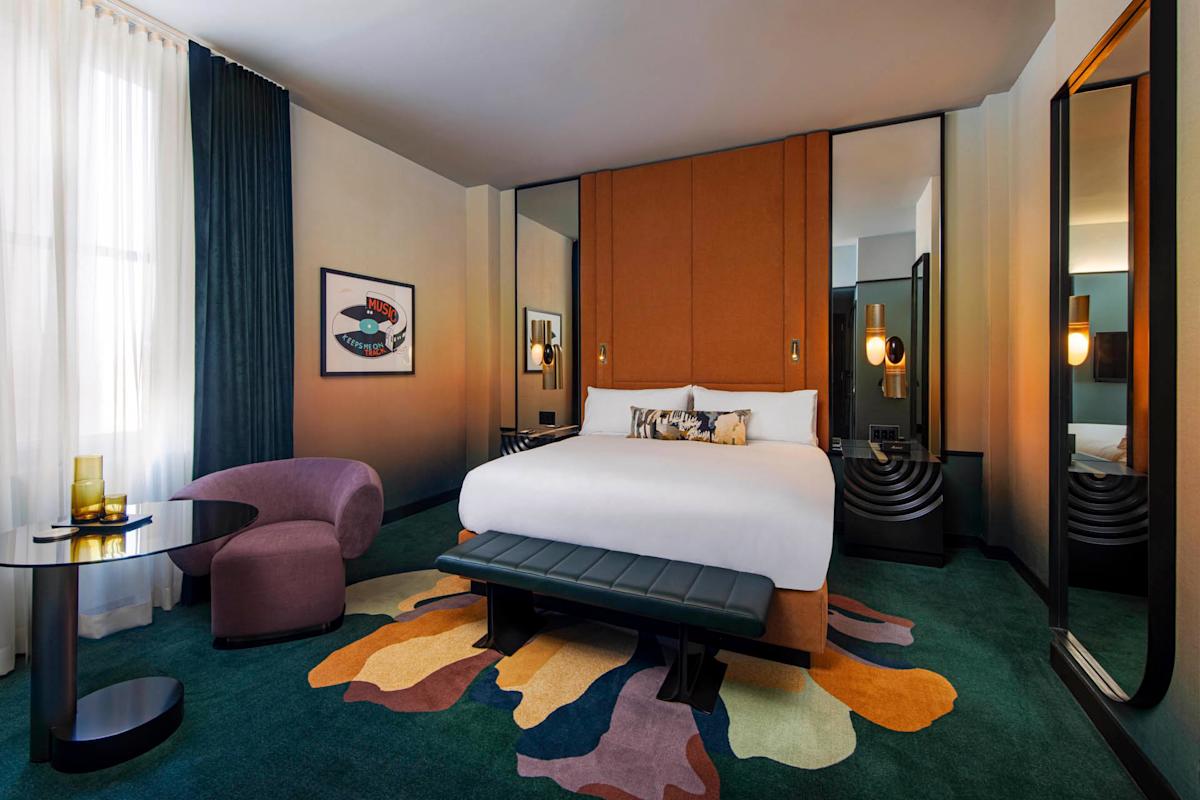 Modern hotel room with a large bed, brown upholstered headboard, colorful abstract rug, purple chair, round glass table, decorative lighting, and tall windows with sheer white curtains and dark drapes.