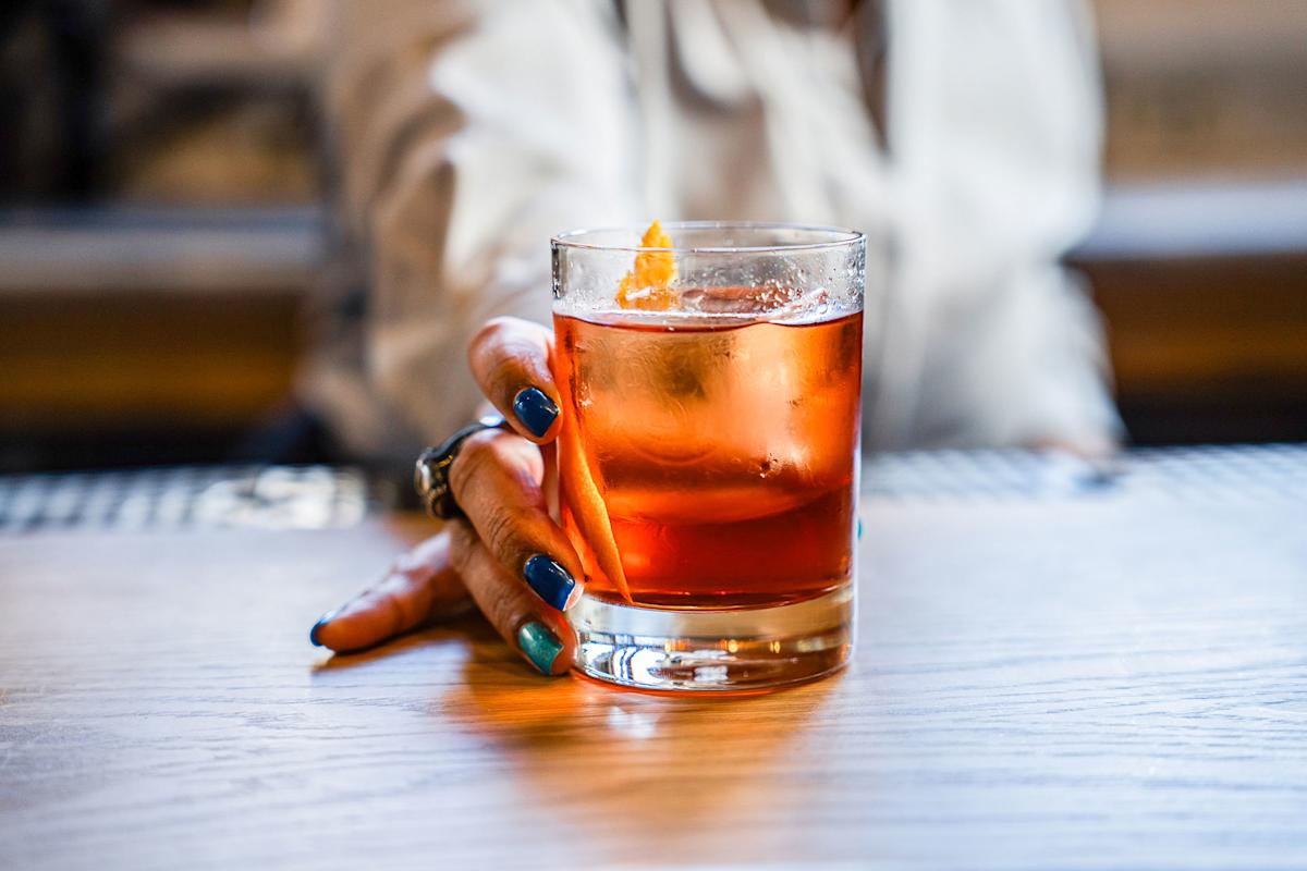A person with blue nail polish holds a glass of orange-colored cocktail with ice and an orange peel garnish, resting it on a wooden table.