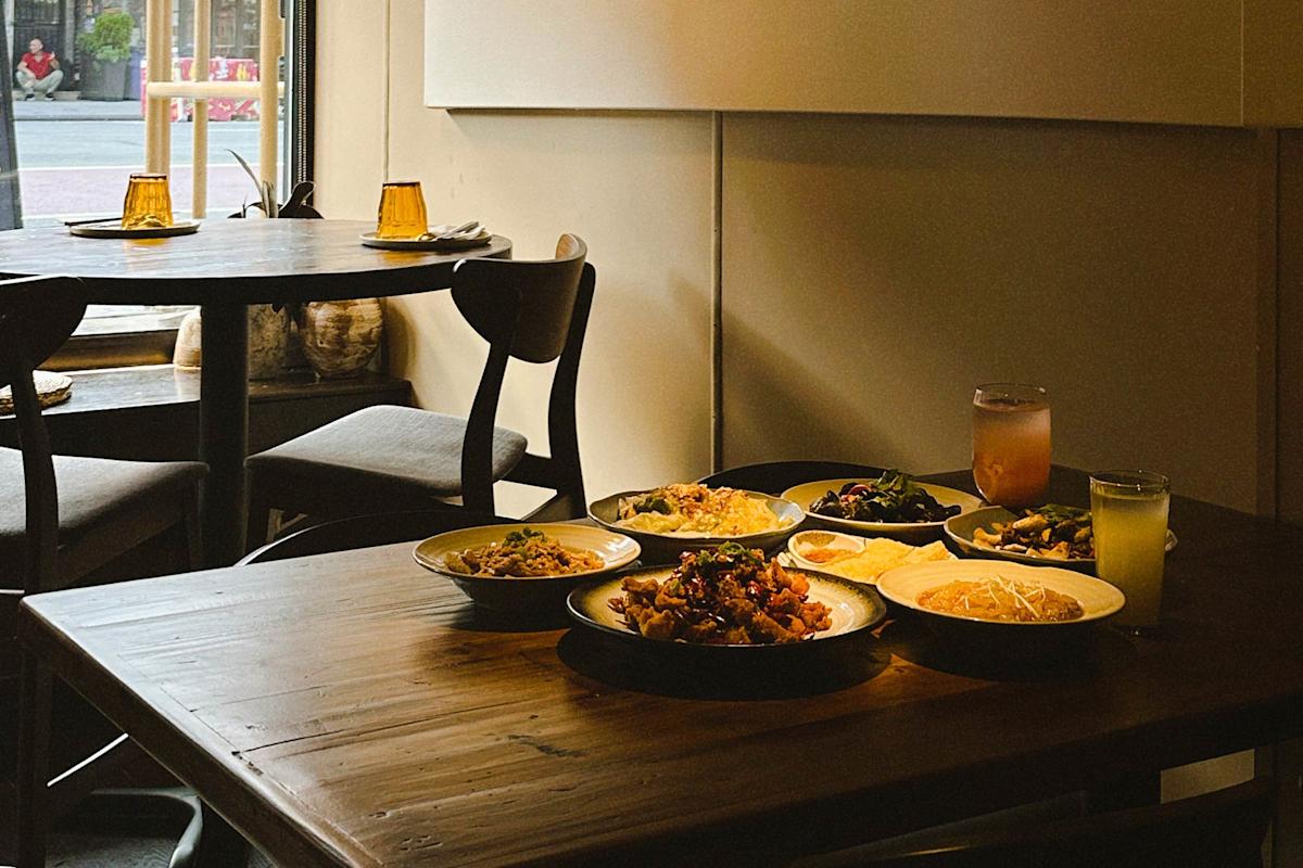 A wooden table set with several plates of food and two drinks in a cozy, dimly lit restaurant near a window. An empty table with chairs is visible in the background.