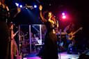A woman in a black dress sings into a microphone on a brightly lit stage, surrounded by band members playing guitar, bass, and drums under colorful lights.