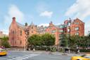 A large red-brick building with multiple gabled roofs and green window frames stands behind trees on a busy city street, with pedestrians walking and yellow taxis passing by.