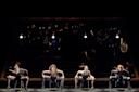 Four female dancers in black outfits perform a synchronized routine on chairs under hanging lights, with a live orchestra playing in the dimly lit background.