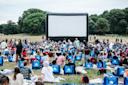 A large crowd of people sits on portable chairs and blankets in a grassy field, facing an inflatable outdoor movie screen. Trees line the background, and the atmosphere is casual and lively.