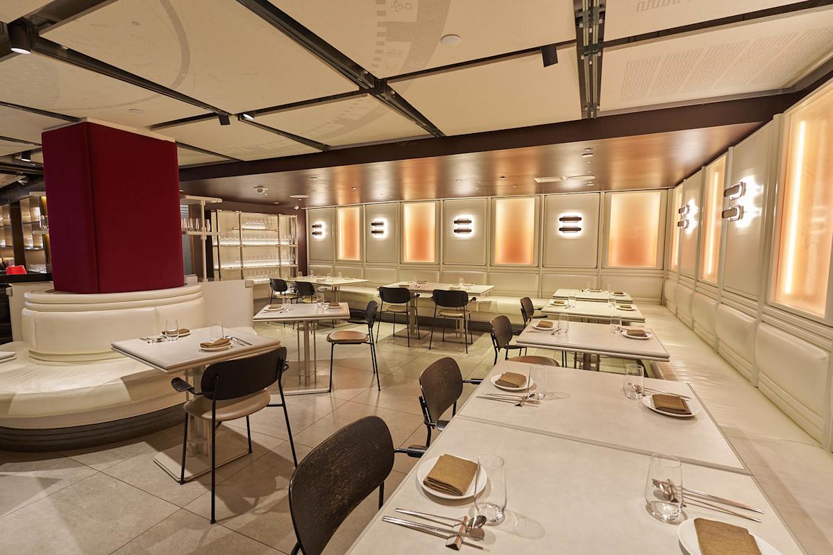 Modern restaurant interior with beige and brown tones, white tables set with plates and napkins, cushioned seating along the wall, soft lighting, and a clean, minimalist design. Room appears empty and ready for guests.