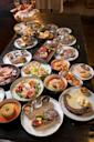 A large table filled with various gourmet dishes, including steaks, seafood platters, pasta, salads, grilled octopus, charcuterie, sides, and wine glasses, all artfully arranged on white and colored plates.