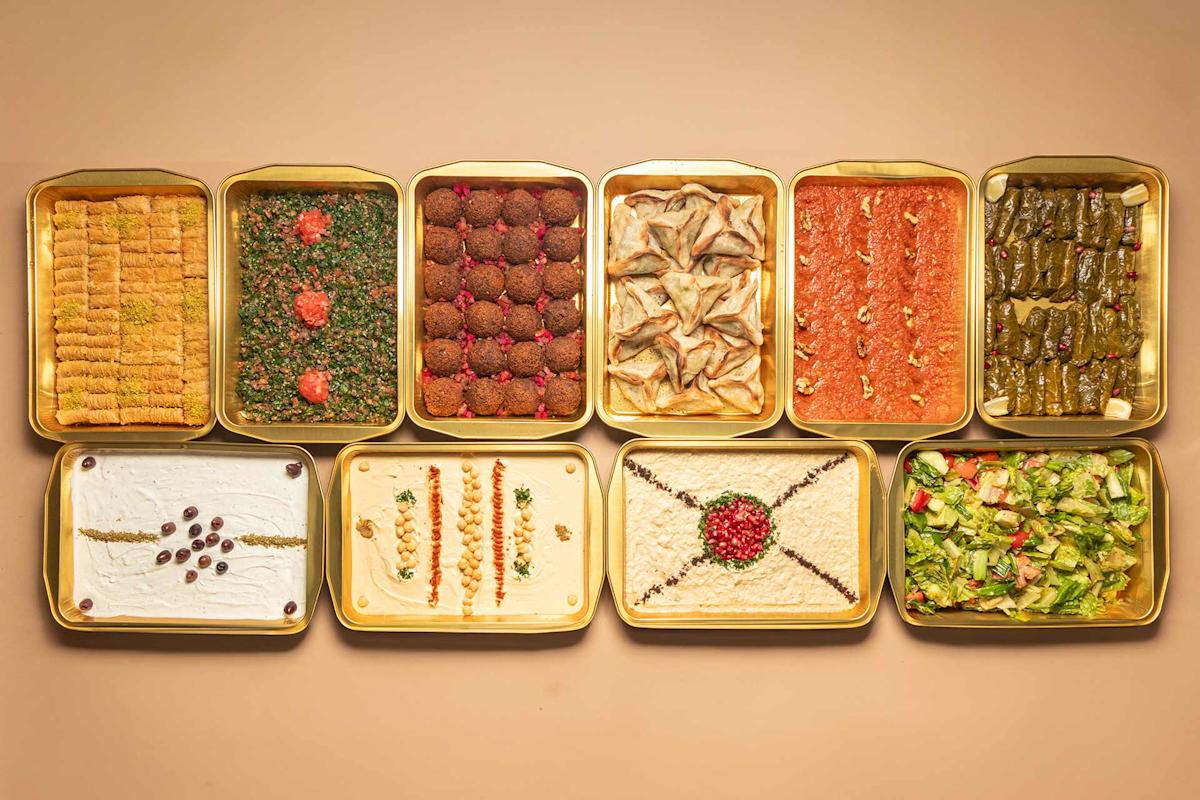 Ten trays of assorted Middle Eastern dishes, including baklava, tabbouleh, falafel, sambousek, red dips, stuffed grape leaves, creamy dips, and salad, arranged neatly on a beige surface.