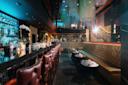Luxurious bar interior with dim lighting. Long bar counter on the left with leather bar stools, and plush booths on the right. Shelves of bottles are backlit in blue light. Elegant decor includes orchids and modern pendant lights.
