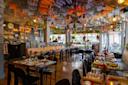 A vibrant restaurant with festive papel picado banners and piñatas hanging from the ceiling, colorful decor, wooden floors, neatly set tables, and plenty of natural light from large windows.