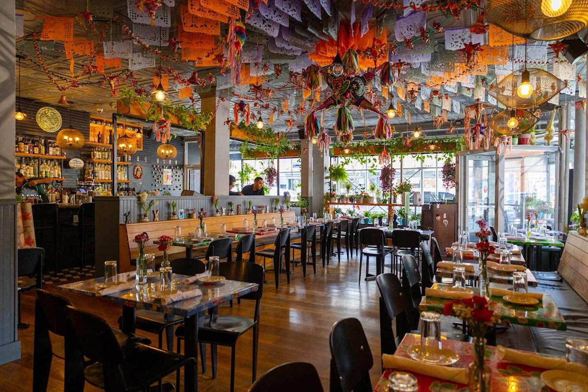 A vibrant restaurant with festive papel picado banners and piñatas hanging from the ceiling, colorful decor, wooden floors, neatly set tables, and plenty of natural light from large windows.
