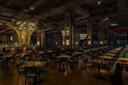 Spacious, modern restaurant interior with dim lighting. Wooden tables and chairs fill the room, with settings for dining. A well-lit bar with tall, artistic light fixtures is in the background. The atmosphere is warm and inviting.