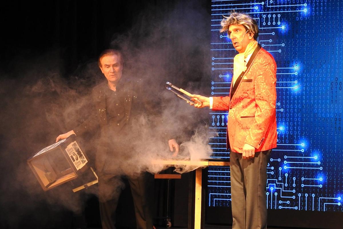 Two magicians perform on stage amid smoke; one, in a black outfit, tips a box, while the other, in a red jacket, holds metal tongs. The backdrop shows a digital circuit pattern.