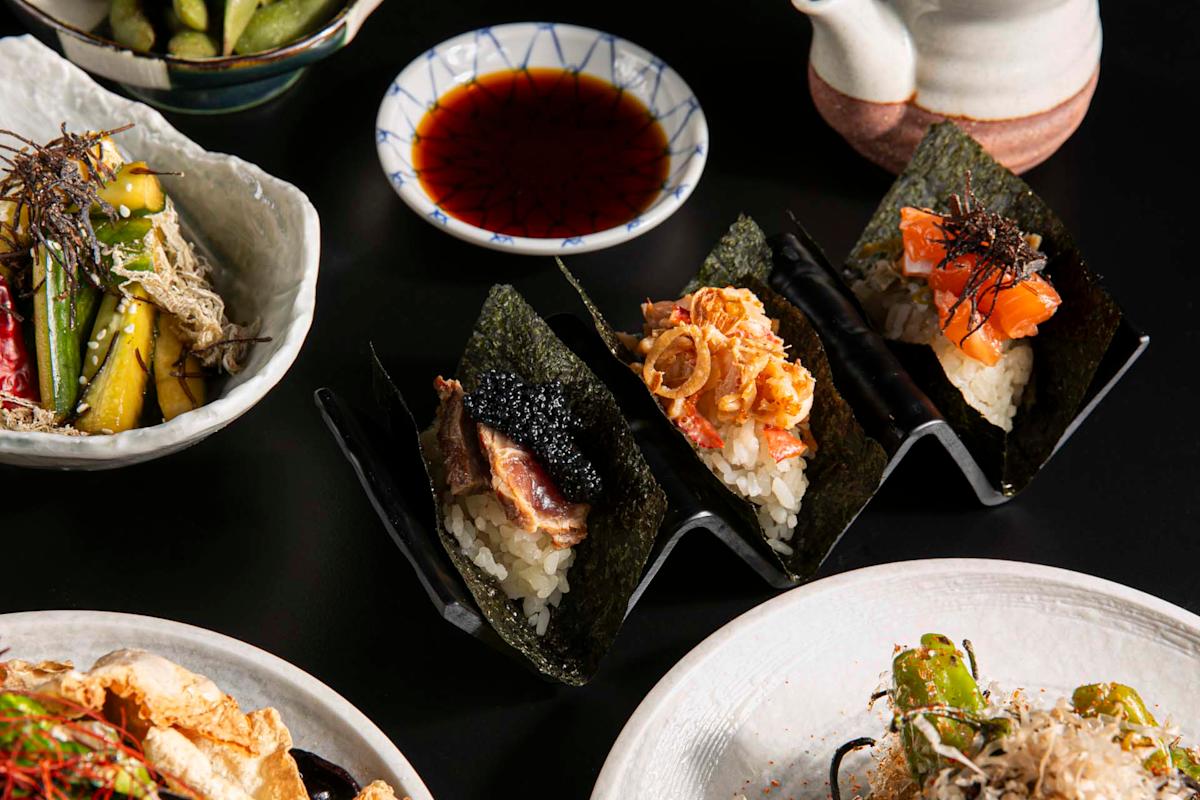 Three hand-rolled sushi cones filled with various fish and toppings are displayed on a sushi holder, surrounded by plates of Japanese dishes and a small bowl of soy sauce.