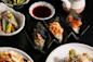 Three hand-rolled sushi cones filled with various fish and toppings are displayed on a sushi holder, surrounded by plates of Japanese dishes and a small bowl of soy sauce.