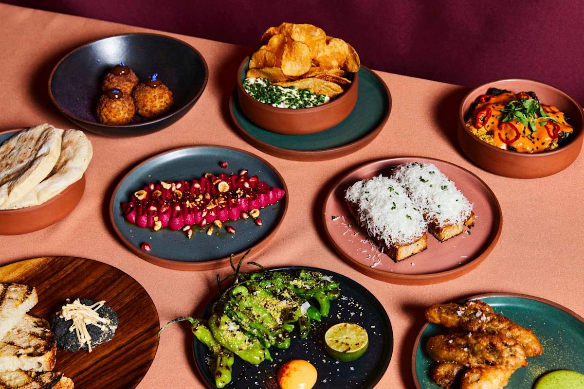 A variety of colorful dishes on a table, including plates of roasted vegetables, seasoned meat, green beans, toast with cheese, sliced bread, potato chips with dip, and falafel. The presentation is vibrant and appetizing.