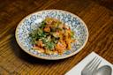 A blue and white patterned bowl filled with a colorful salad of herbs, sliced vegetables, and a red spicy sauce sits on a wooden table next to a fork and spoon on a napkin.