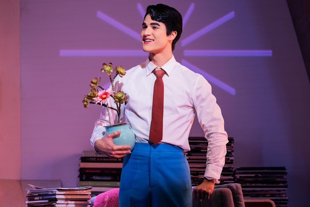 A young man in a white shirt, red tie, and blue pants smiles while holding a potted plant. He stands indoors in front of a stack of magazines and a purple light pattern on the wall behind him.