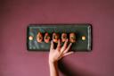 A hand reaches for a piece of sushi on a rectangular black plate. The plate holds five pieces of sushi, garnished with vibrant toppings. The background is a deep burgundy color.