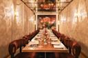 A long dining table with elegant place settings, surrounded by plush red chairs, sits in a warmly lit room with floral artwork and mirrored ceiling panels, creating a cozy, upscale atmosphere.