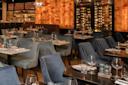 Restaurant interior with tables set for dining, featuring blue and gray chairs. Walls display warm wooden panels and a wine rack. The ambiance is elegant, with neatly arranged glassware and cutlery on the tables. Hanging lights add a soft glow.