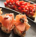 Close-up of bruschetta appetizers topped with creamy cheese, thin slices of prosciutto, and shaved truffle, with another type of bruschetta topped with diced tomatoes in the background.