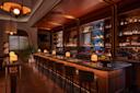 A stylish, dimly lit bar with a long marble counter, high-backed stools, shelves of liquor bottles, glowing lamps, and warm wood accents, creating an inviting and elegant atmosphere.