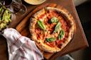 A wood-fired Margherita pizza topped with tomato sauce, melted cheese, and fresh basil leaves sits on a wooden board next to a glass of red wine and a white napkin with red stripes.
