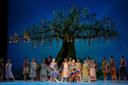 A group of performers in colorful costumes dance and pose on stage before a large, decorated tree with hanging ornaments and ladders, set against a blue backdrop.