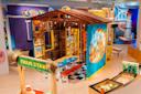 A colorful indoor play area features a wooden playhouse decorated with bright artwork, picnic tables, a “Farm Stand” sign, play food, and various kid-friendly toys and activities. The scene is lively and inviting for children.