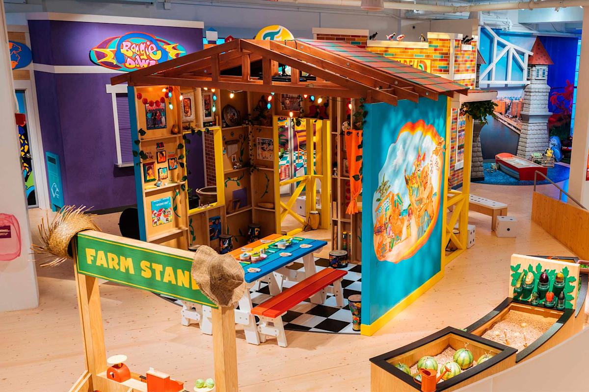A colorful indoor play area features a wooden playhouse decorated with bright artwork, picnic tables, a “Farm Stand” sign, play food, and various kid-friendly toys and activities. The scene is lively and inviting for children.
