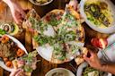 A group of people share slices of arugula-topped pizza around a wooden table filled with various dishes, including grilled fish, roasted vegetables, and colorful drinks.