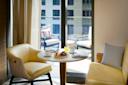 A cozy dining area with two yellow chairs and a round table set for breakfast, featuring fruit, coffee, and juice. The table is next to a large window overlooking a balcony with cushioned seating and a view of a building outside.