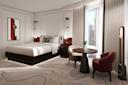 A modern hotel room with a large bed, decorative pillows, abstract wall art, a round table with two chairs, floor-to-ceiling windows with city views, and carpet with a wavy pattern.