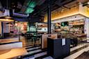 Modern restaurant interior with black and white tiled floor, wooden booths, bar seating, shelves of bottles, industrial ceiling, and colorful graffiti art on the wall in the background.