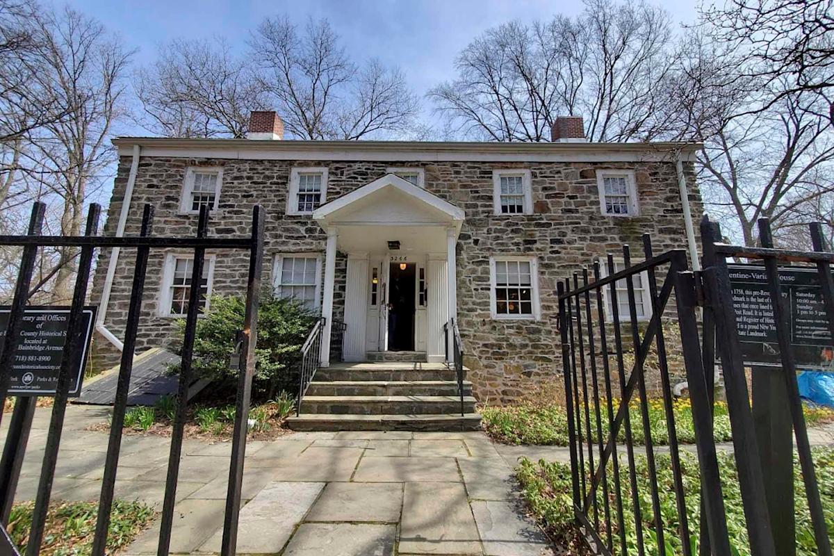 An old stone house with white-framed windows and a central door sits behind a black wrought iron fence. Steps lead up to the entrance under a small portico. Bare trees and overcast skies are visible in the background.