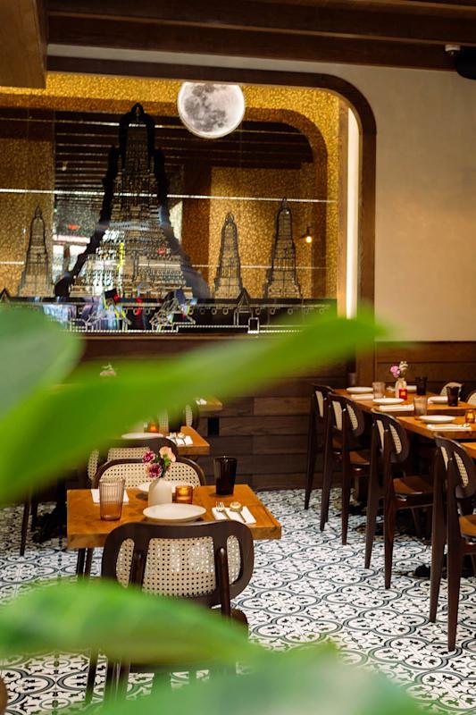 A cozy restaurant with patterned tile floors, wooden tables and chairs, floral centerpieces, and a mural of a temple on the back wall. Green plant leaves are visible in the foreground.